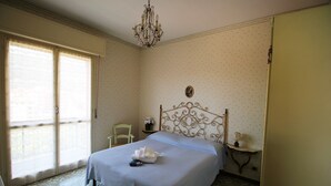 A neatly made bed with a blue bedspread and white linens, a chandelier, a small bedside table with a lamp, and a chair. - House Adelin (Andora)