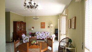 A dining area with a round wooden table, floral chairs, a chandelier, a cabinet, and a sofa with a patterned cover. - House Adelin (Andora)