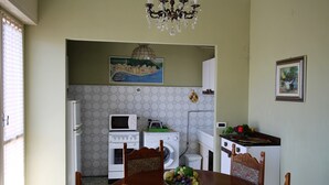 A dining area with a table set for four, a chandelier, and a kitchen in the background with appliances and a painting. - House Adelin (Andora)