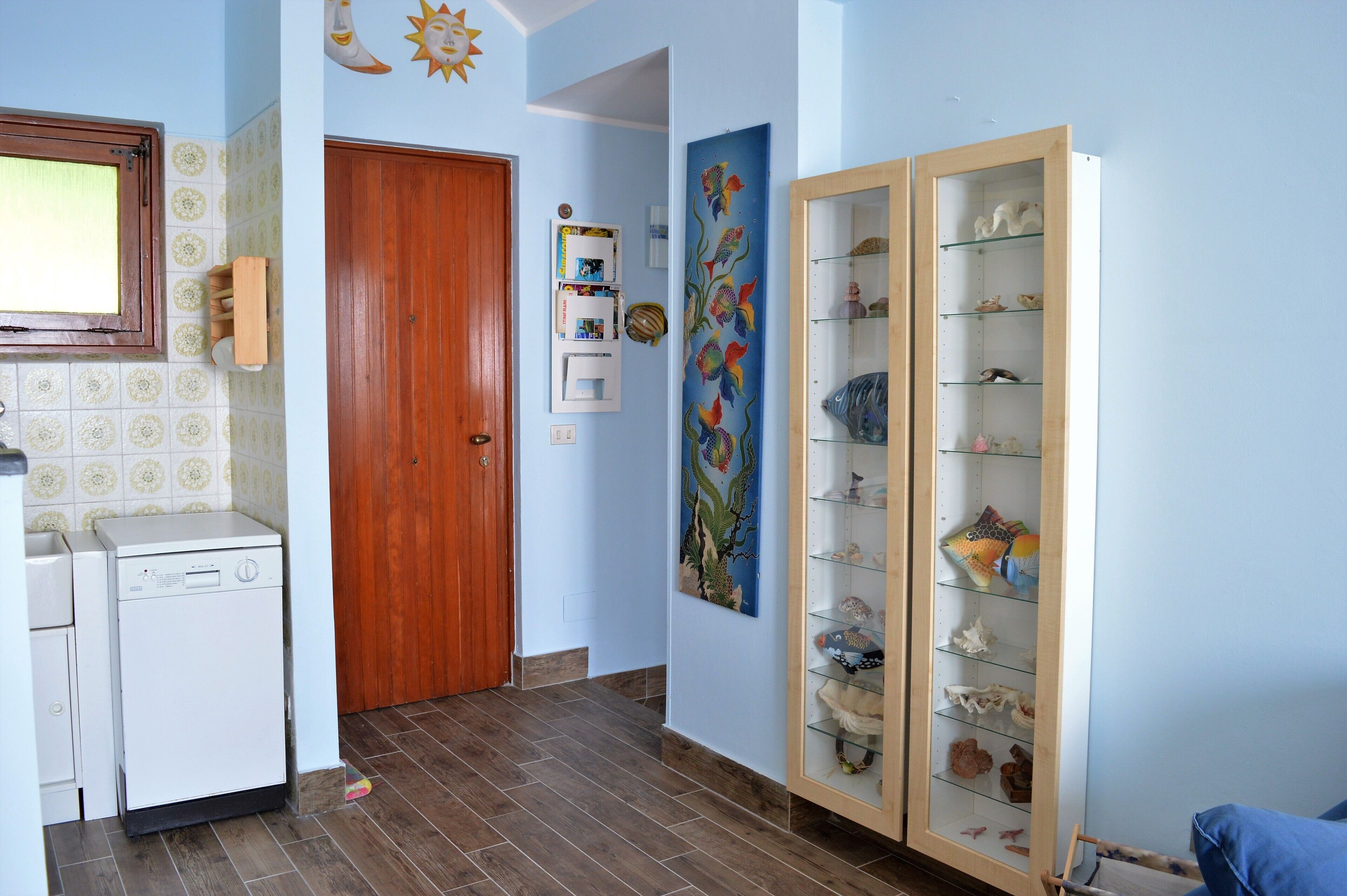 Un pasillo con una puerta de madera, un electrodoméstico blanco, un estante montado en la pared y un aparador de vidrio con objetos decorativos.