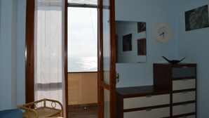 A room with a wooden door leading to a balcony, a small wooden chair, a dresser with drawers, a wall clock, and a framed picture. - House Agave (Moneglia)