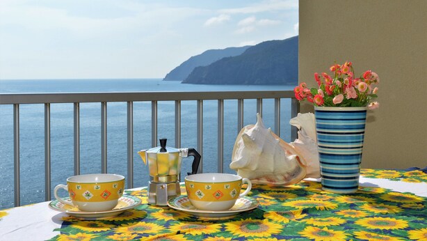 A table with two yellow cups and a coffee pot, a potted plant, and a seashell on a balcony overlooking the sea. - House Agave (Moneglia)