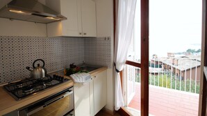 Une cuisine équipée d’une cuisinière à gaz, d’une bouilloire et d’un évier. Il y a une fenêtre avec des rideaux, et la vue donne sur les bâtiments environnants.