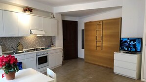 A modern kitchen with white cabinets, a tiled backsplash, and a stove. There's a dining table with chairs and a potted plant. A TV is on a white cabinet, and a wooden door with a ladder is visible. - House Orsa Maggiore (Bordighera)