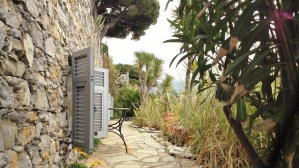 A stone pathway leading to a building with a metal shutter door, surrounded by various plants and trees. - House Roberta (Recco)