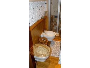 A bathroom with a stone wall, a wooden bench, a toilet with a decorative bowl, and a shower area. - House Limone (Recco)