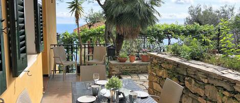 Un espace extérieur aménagé pour les repas, avec une table dressée pour quatre personnes, entourée de chaises en osier, sur un balcon doté d’un mur en pierre et de plantes en pot.