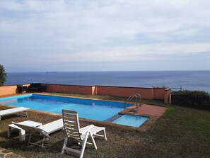 Una piscina con vista al mar, rodeada de sillas de descanso y un muro de piedra.