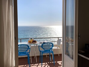 Un balcon avec une table dressée pour deux personnes, agrémentée d’un plateau de fruits et d’un livre, donnant sur une mer d’un bleu limpide.