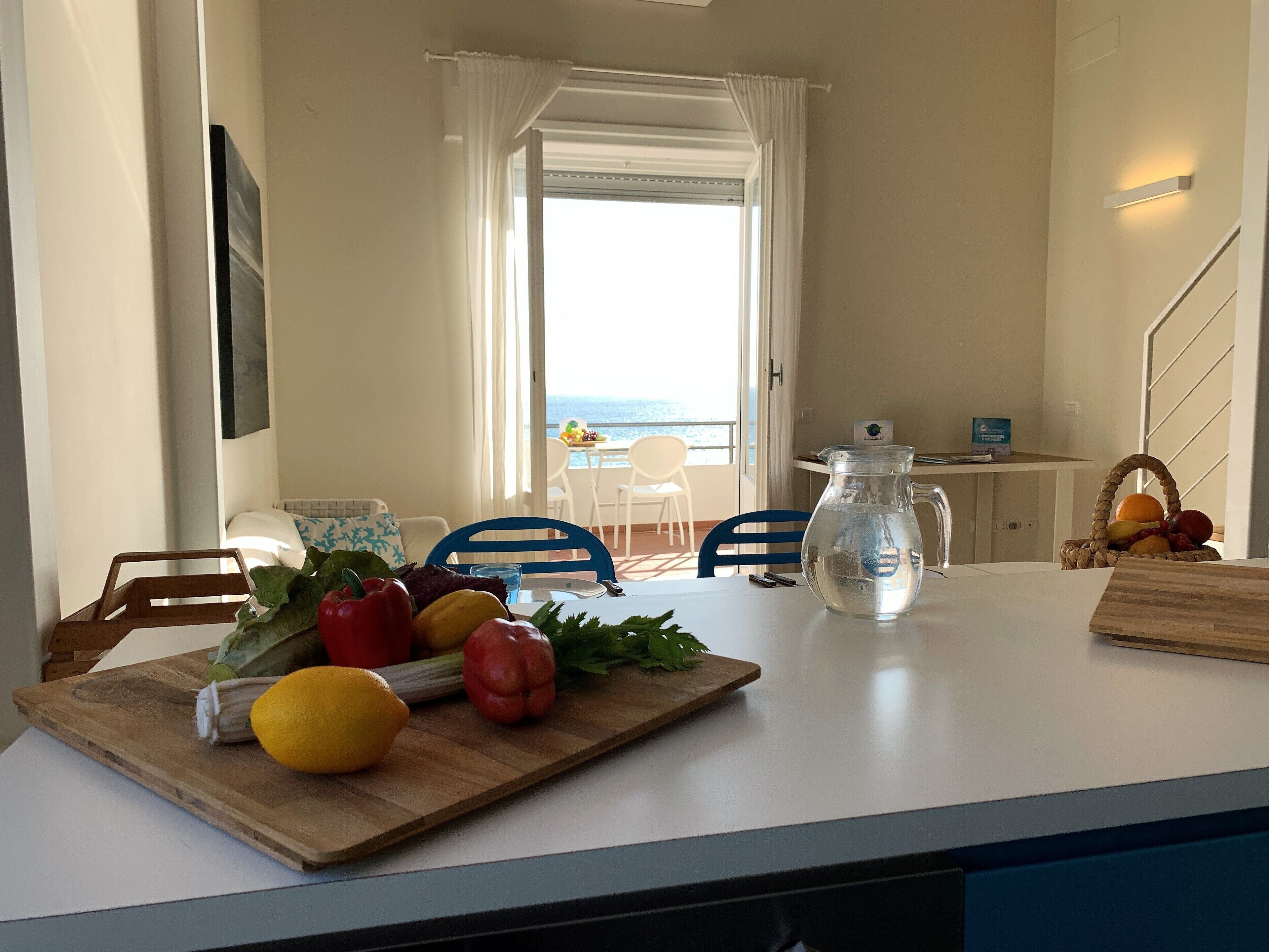 A kitchen with a wooden cutting board, fresh vegetables, a pitcher, and a view of the sea through a window.