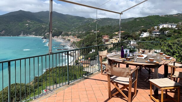 Terraço com vista para o mar e montanhas, mobiliário de madeira e uma mesa com garrafa e tigelas.