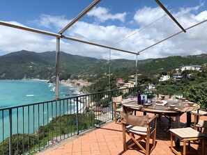 Une terrasse offrant une vue sur la mer et les montagnes, avec des meubles en bois et une table sur laquelle se trouvent une bouteille et des bols.