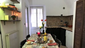 A dining area with a table set for two, a radiator, a shelf with books and a candle, and a view of the kitchen. - House Cornelia (Pietra Ligure)