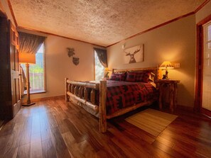 Een slaapkamer met een houten bed, een houten hoofdeinde, een raam met zonwering, een lamp op een nachtkastje en een aan de muur bevestigd decoratie van een hertenkop.