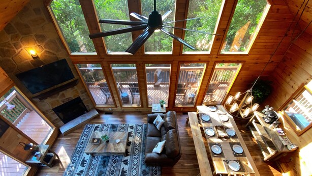 A spacious living area with a large sofa, a coffee table, and a dining table set for four. The room features wooden floors, a stone fireplace, and large windows offering a view of the outdoors. - Stunning Pigeon Forge Pet-Friendly A-Frame Cabin (Sevierville)