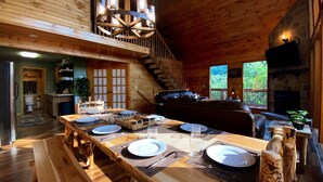 A wooden interior with a dining table set for four, a staircase, a fireplace, and a view of greenery outside. - Stunning Pigeon Forge Pet-Friendly A-Frame Cabin (Sevierville)