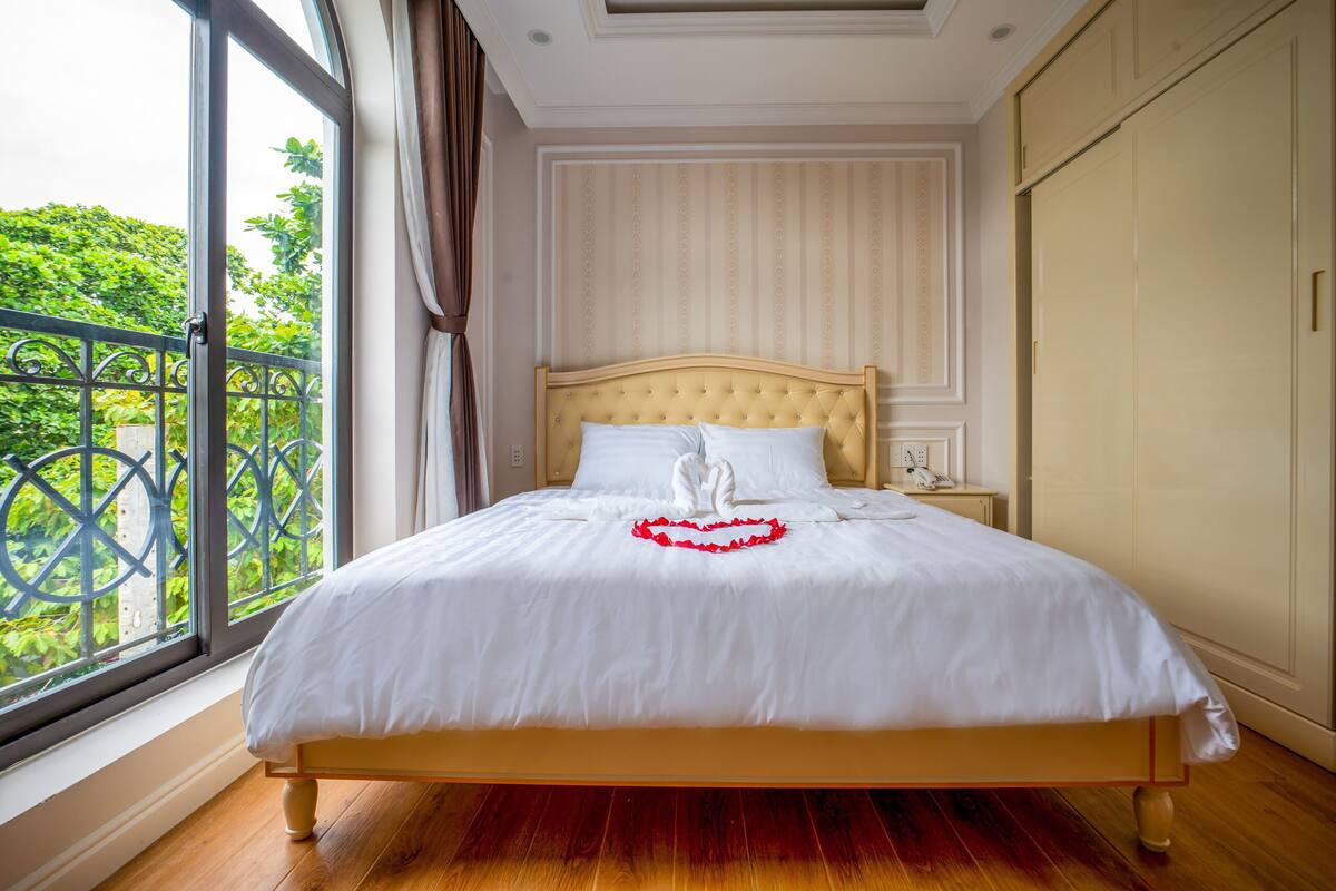 A neatly made bed with white linens and a red heart decoration, a bedside table with a lamp, and a view of greenery through a window with a metal railing.