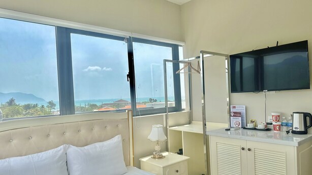 Un cuarto de hotel con cama, una mesita de noche, un televisor montado en la pared y una ventana que ofrece vistas al mar y a edificios.