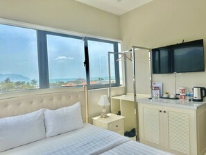 Un cuarto de hotel con cama, una mesita de noche, un televisor montado en la pared y una ventana que ofrece vistas al mar y a edificios.