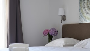 Ein ordentlich bezogenes Bett mit weißen Leinen, eine Vase mit rosa und lila Blumen und eine Wandlampe.