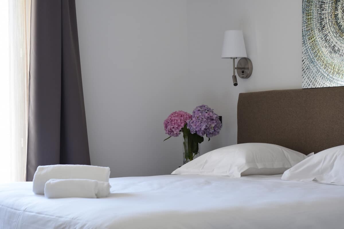 Una cama bien hecha con sábanas blancas, un jarrón con flores rosas y moradas, y una lámpara fijada a la pared.