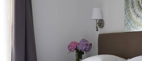 Un lit bien fait, avec du linge de lit blanc, un vase rempli de fleurs roses et violettes, et une lampe fixée au mur.
