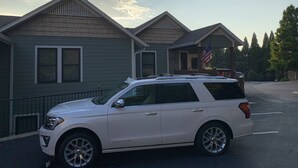 Een witte SUV geparkeerd voor een rij huizen, met een vlag aan de rechterkant.
