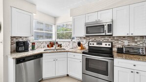 Una cocina moderna con armarios blancos, electrodomésticos de acero inoxidable, lavavajillas, microondas, tostadora y una ventana con persianas.