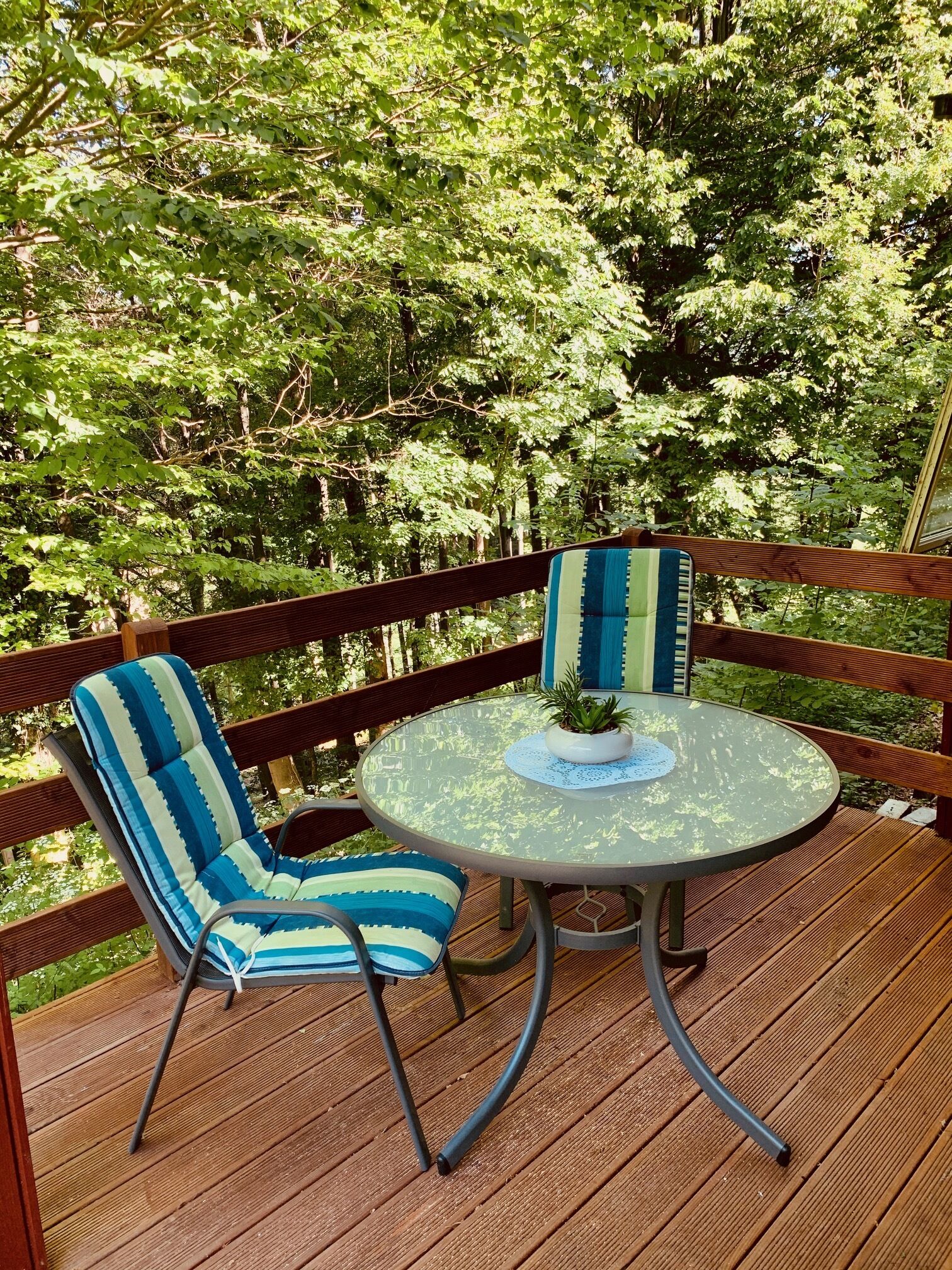 Une terrasse avec deux chaises à rayures et une table ronde, entourée d’une rambarde en bois et d’un feuillage verdoyant et luxuriant.