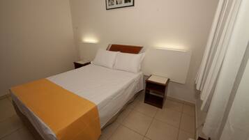 Una cama bien hecha con sábanas blancas y una manta amarilla, dos mesitas de noche con lámparas, y un cuadro en la pared.