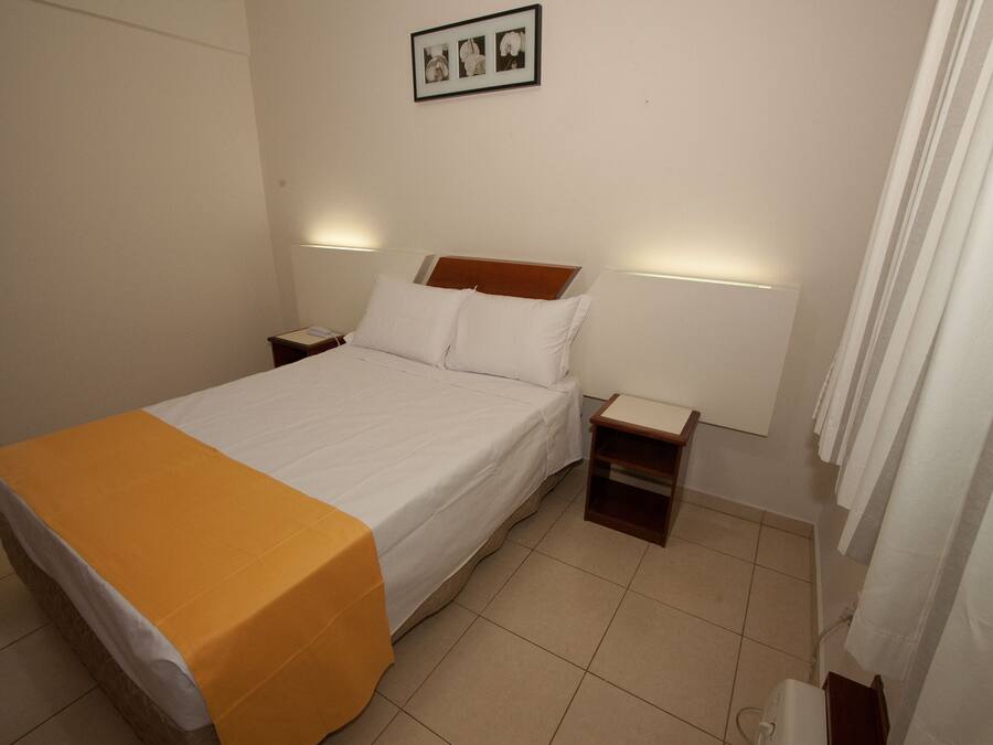 Una cama bien hecha con sábanas blancas y una manta amarilla, dos mesitas de noche con lámparas, y un cuadro en la pared.