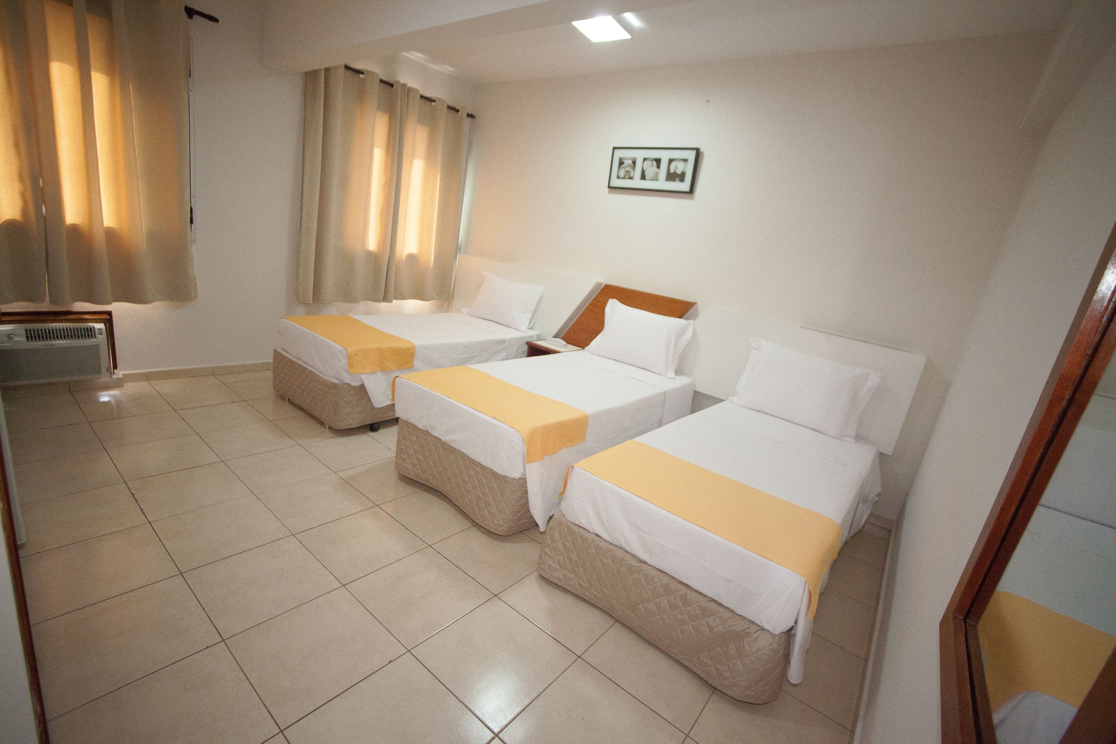 Una habitación de hotel con tres camas individuales, cada una con un colchón blanco y una colcha amarilla. Hay una mesita de noche pequeña con una lámpara y una ventana con cortinas.
