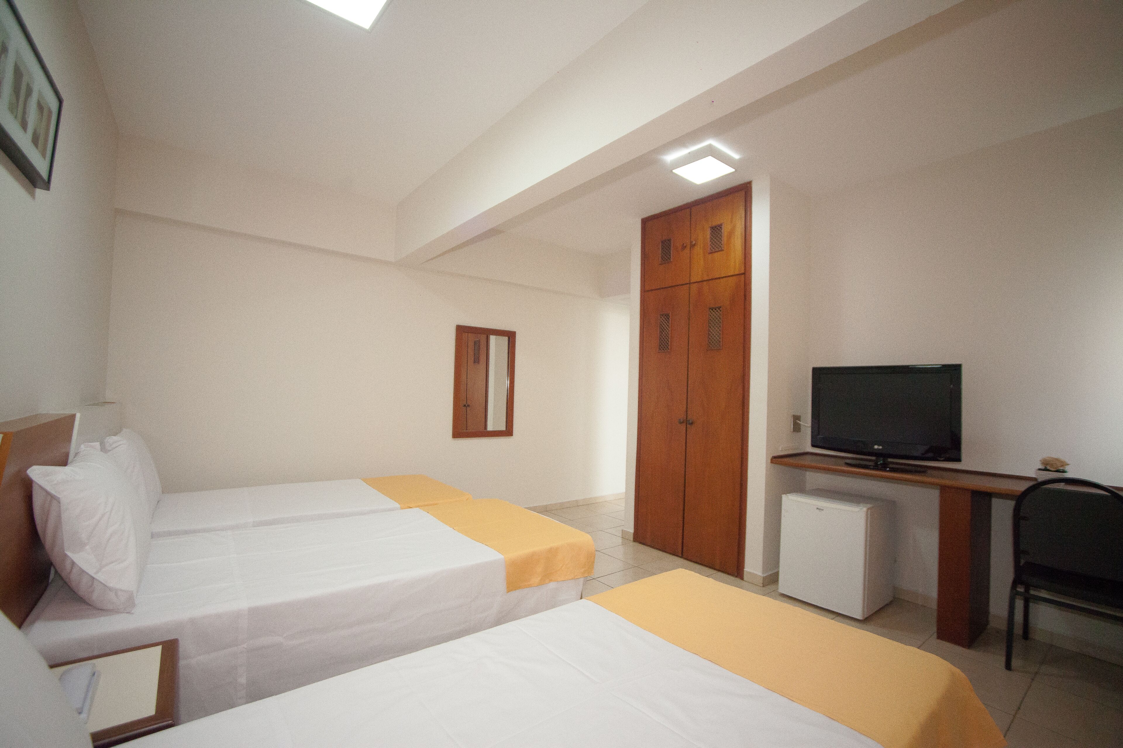 A hotel room with two single beds, a television, a small refrigerator, and a wooden wardrobe.
