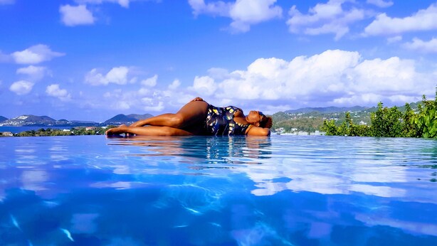 Eine Person entspannt in einem klaren, blauen Infinity-Pool mit Blick auf Berge und Grün im Hintergrund.