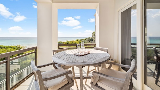 A round wooden table with four chairs, a glass pitcher, and a wine glass on a balcony overlooking the ocean.