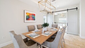 A dining area with a wooden table set for six, surrounded by grey upholstered chairs. A modern chandelier hangs above, and a colorful painting adorns the wall.