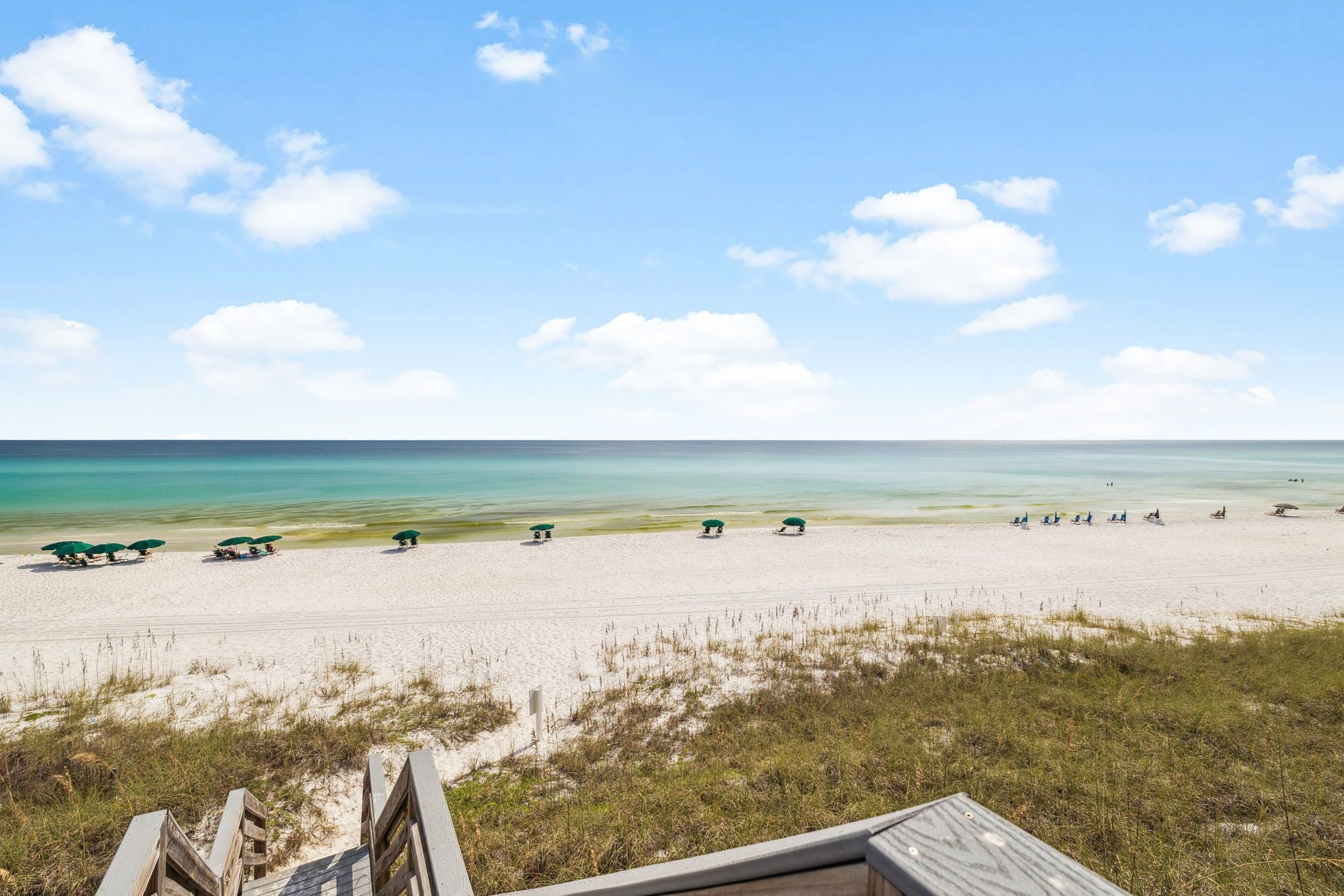 A beach with a wooden walkway leading to the shore, a row of green umbrellas, and a clear view of the ocean.