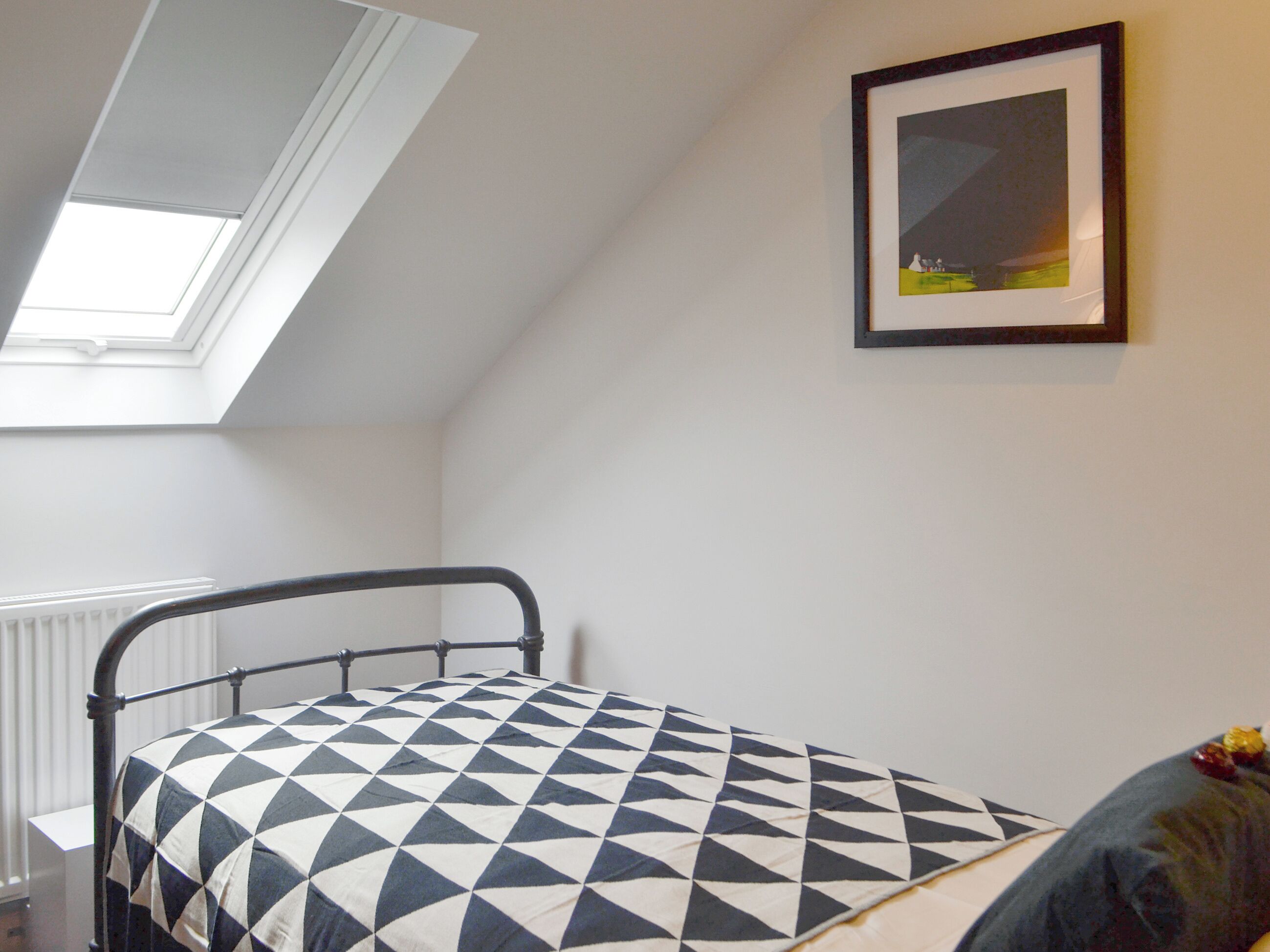 Une chambre avec un puits de lumière, un lit recouvert d’une couvre-lit à motifs géométriques, un tableau encadré au mur et un radiateur.