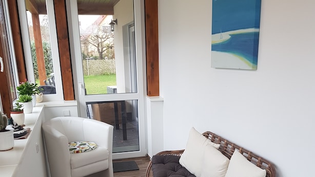 A cozy room with a white sofa, a wicker chair with cushions, a small white table, and a view of a garden through a glass door. - Blooming vacation home at Scharmützelsee (Wendisch Rietz)