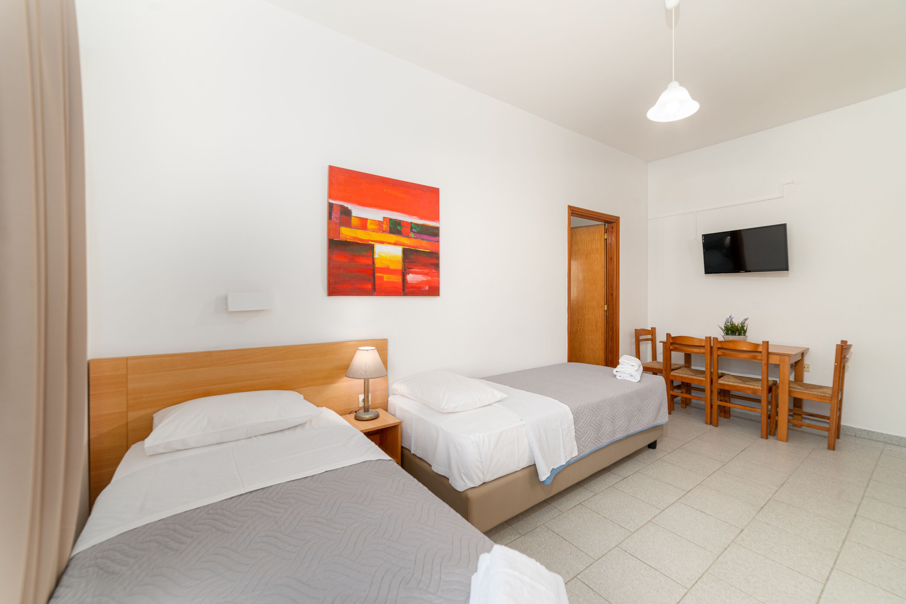 A hotel room with two single beds, a desk with chairs, a television, and a painting on the wall.