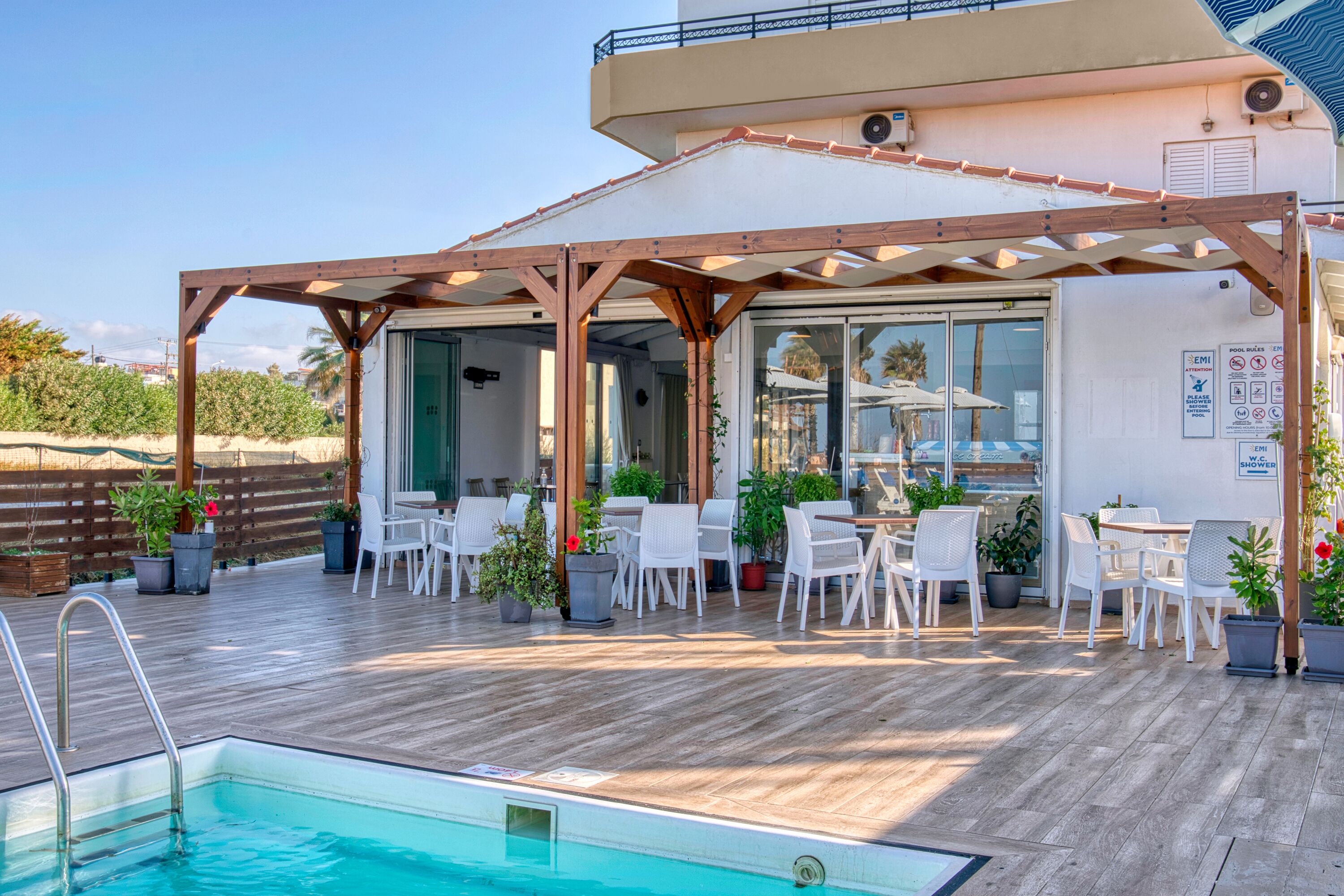 A poolside area with a wooden deck, white chairs, and tables under a pergola.