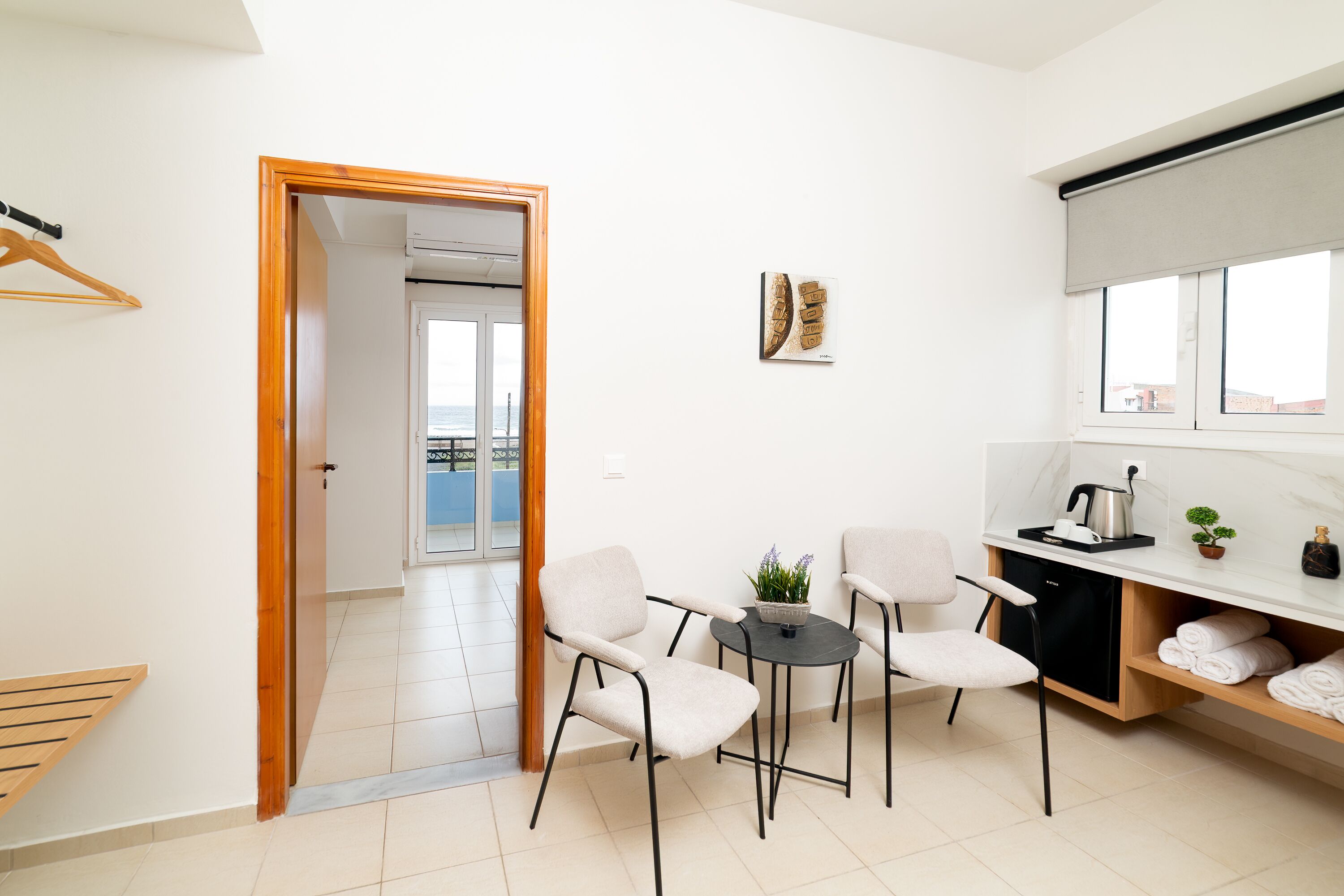 A modern, well-lit room with a wooden door leading to a balcony, a small table with chairs, a kitchen area with a sink and countertop, and a view of the city through the window.