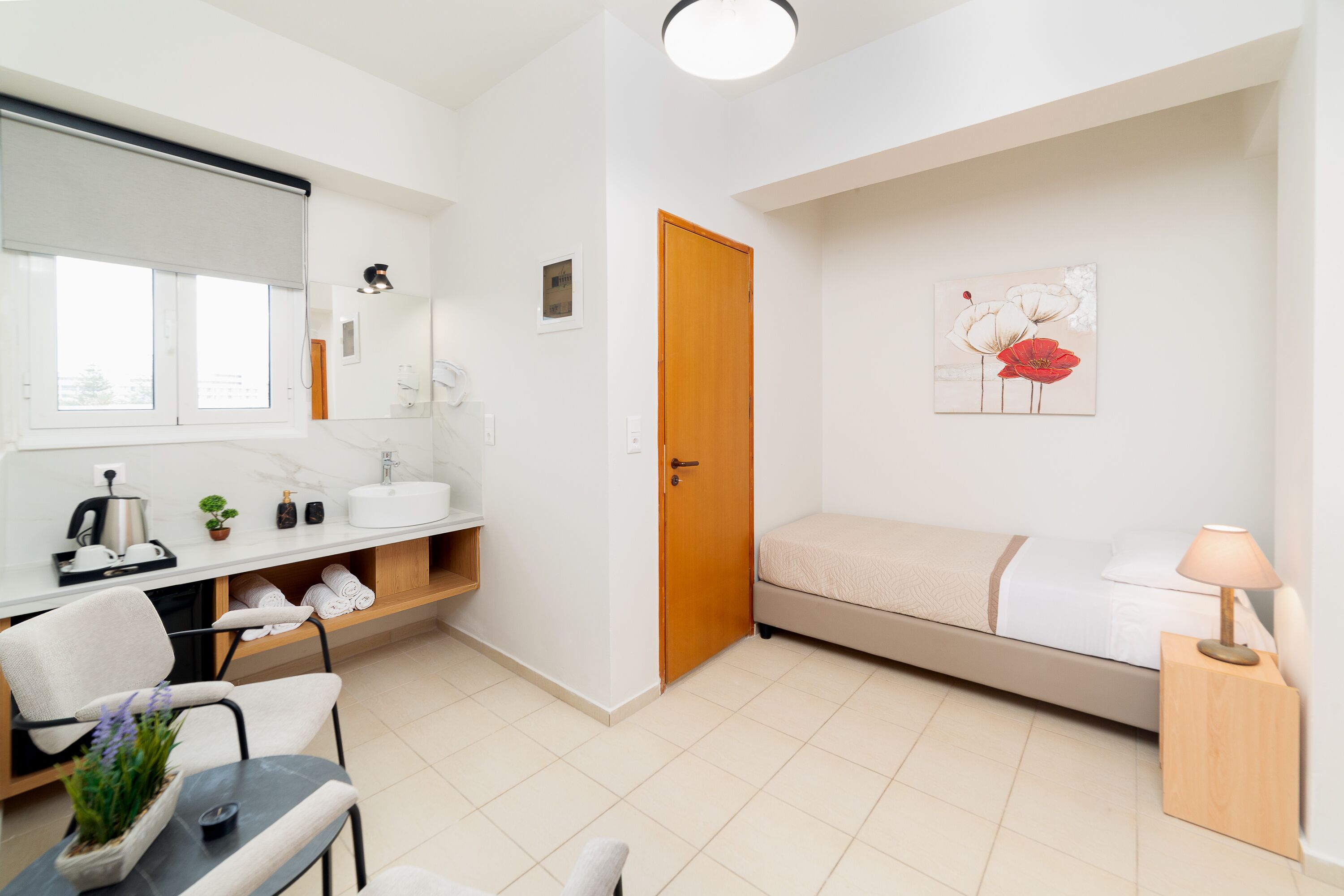 A compact hotel room with a bed, a small desk with a chair, a bathroom with a sink and mirror, and a window with a view.