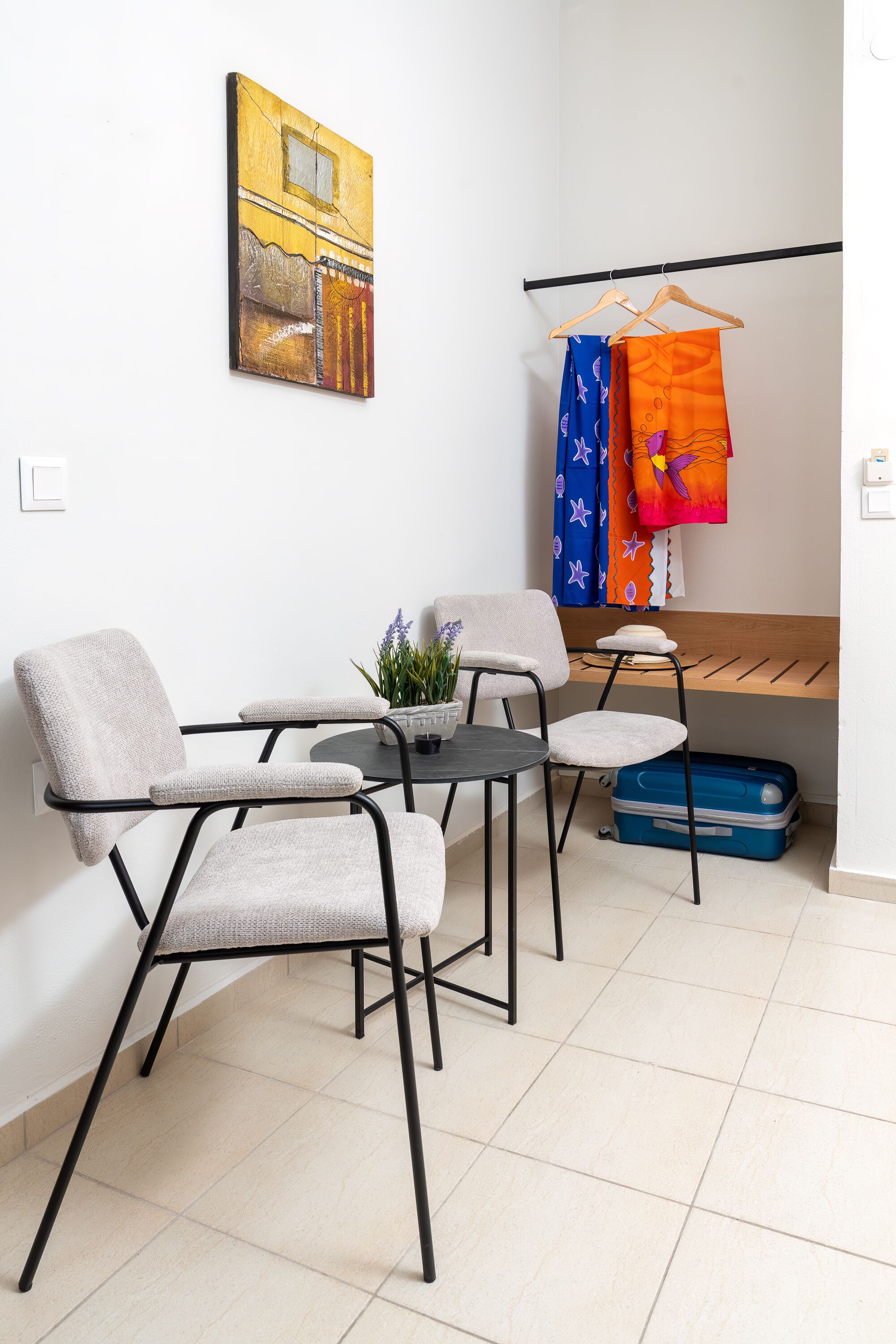 A room with a bed, a desk, two chairs, a small table with a plant, and a hanging clothes rack with colorful garments.