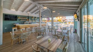 A spacious café with wooden tables and chairs, a bar area with shelves of bottles, and large windows offering a view of the outside. - Emi Seaside (Malevizi)