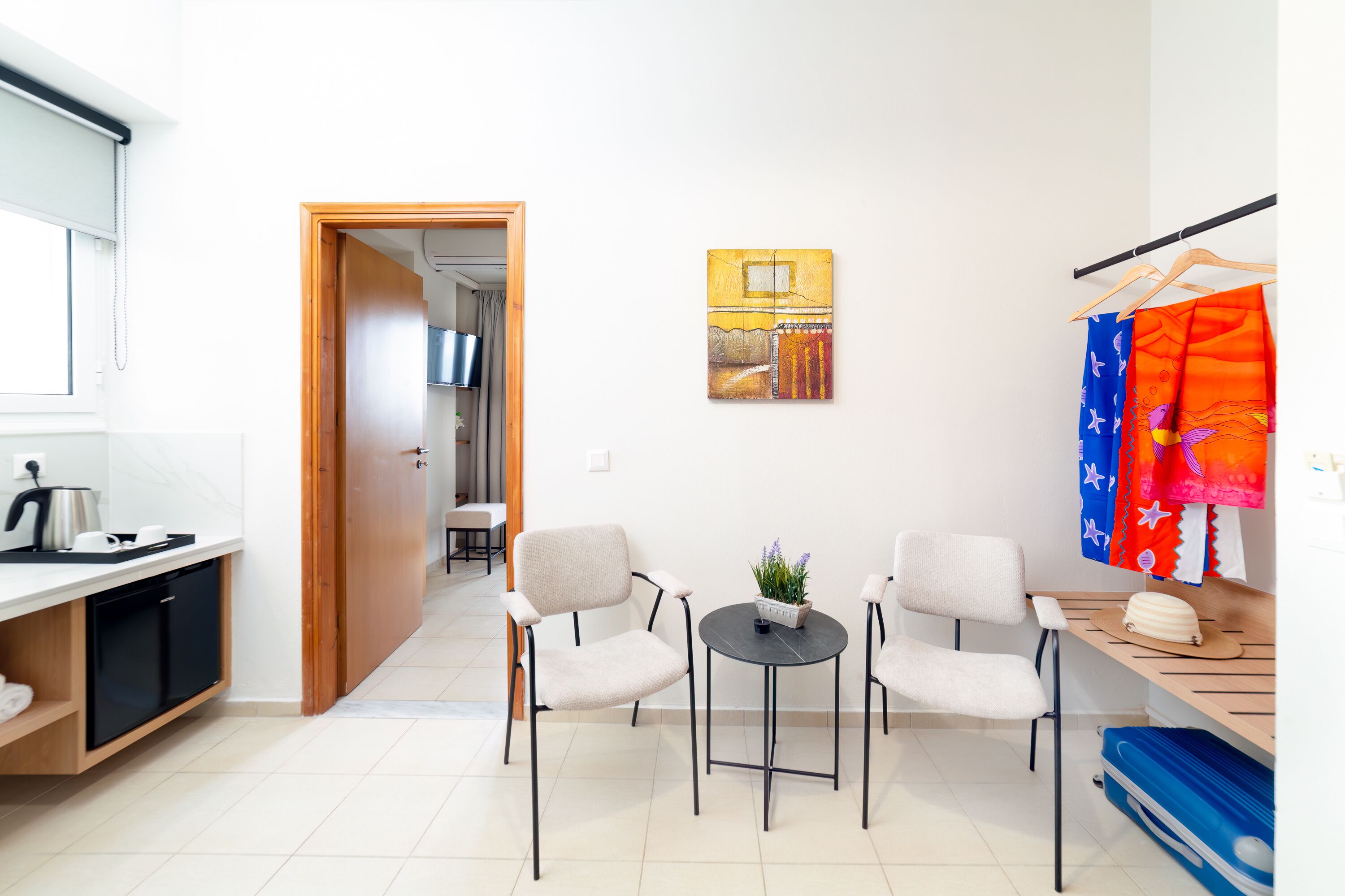 Eine moderne Küche mit Spüle, Wasserkocher und kleinem Kühlschrank. Ein Essbereich mit zwei Stühlen und einem kleinen Tisch ist ebenfalls vorhanden. An der Wand hängt ein Bild, und ein Kleiderständer mit bunten Kleidungsstücken ist sichtbar.