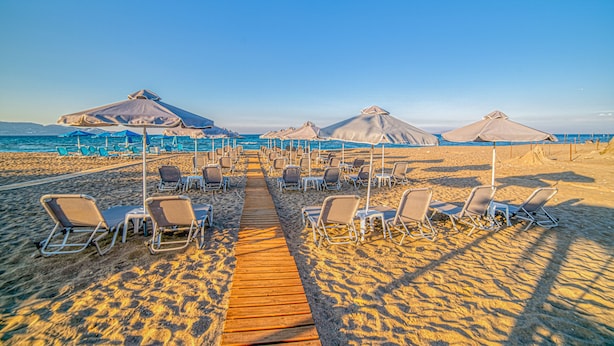 A beach with rows of sun loungers and umbrellas, a wooden walkway, and the ocean in the background. - Emi Seaside (Malevizi)