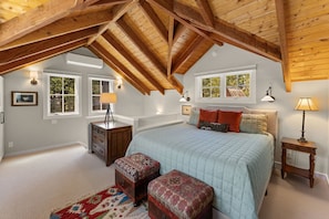 Une chambre mansardée avec un plafond en pente, un lit recouvert d’une courtepointe, deux tables de chevet avec des lampes, une commode et un tapis à motifs.