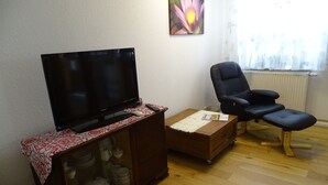 Een woonkamer met een flatscreen televisie op een houten kast, een zwarte bureaustoel, een klein houten tafeltje en een radiator.