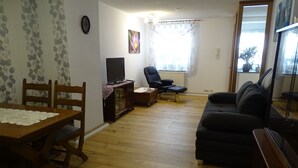 Een woonkamer met een houten eettafel, twee houten stoelen, een zwarte bank, een zwarte fauteuil, een televisie en een radiator.